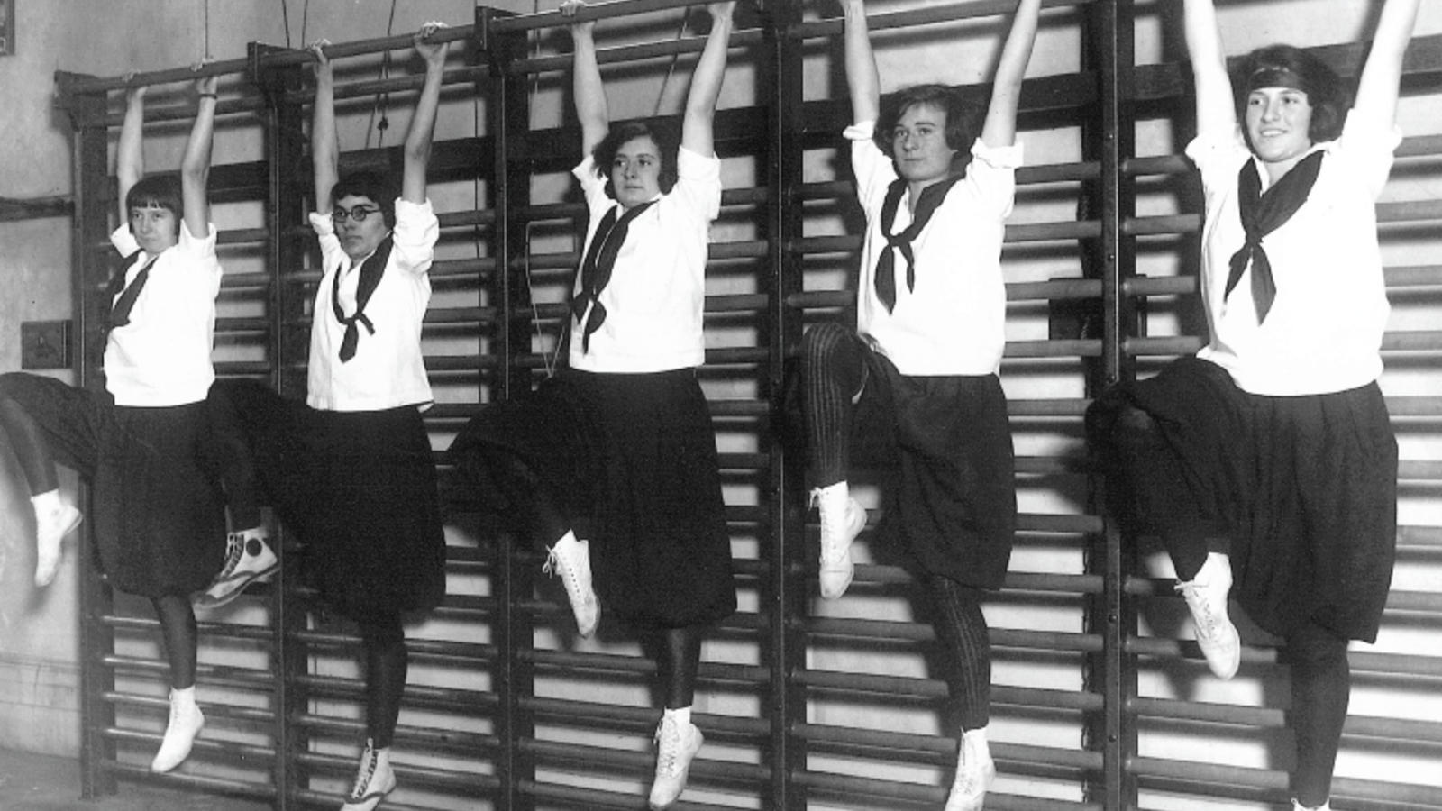 5 women dressed in old-fashioned exercise outfits hang from a bar in gym class